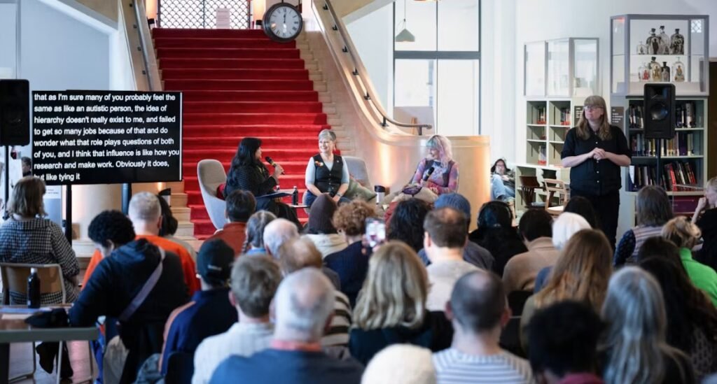 A seated audience in  front of a three person panel. A BSL translator is standing on the stage next to them.
