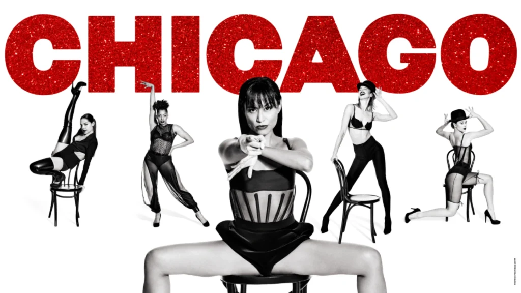 Five dancers in black and white sit under the red glittery Chicago sign using chairs as props