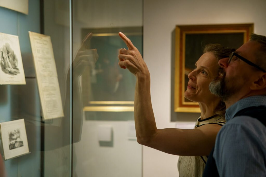 A woman in a white vest points at a paper artefact in a glass cabinet. Beside her is a man in a blue shirt, looking where she's pointing.